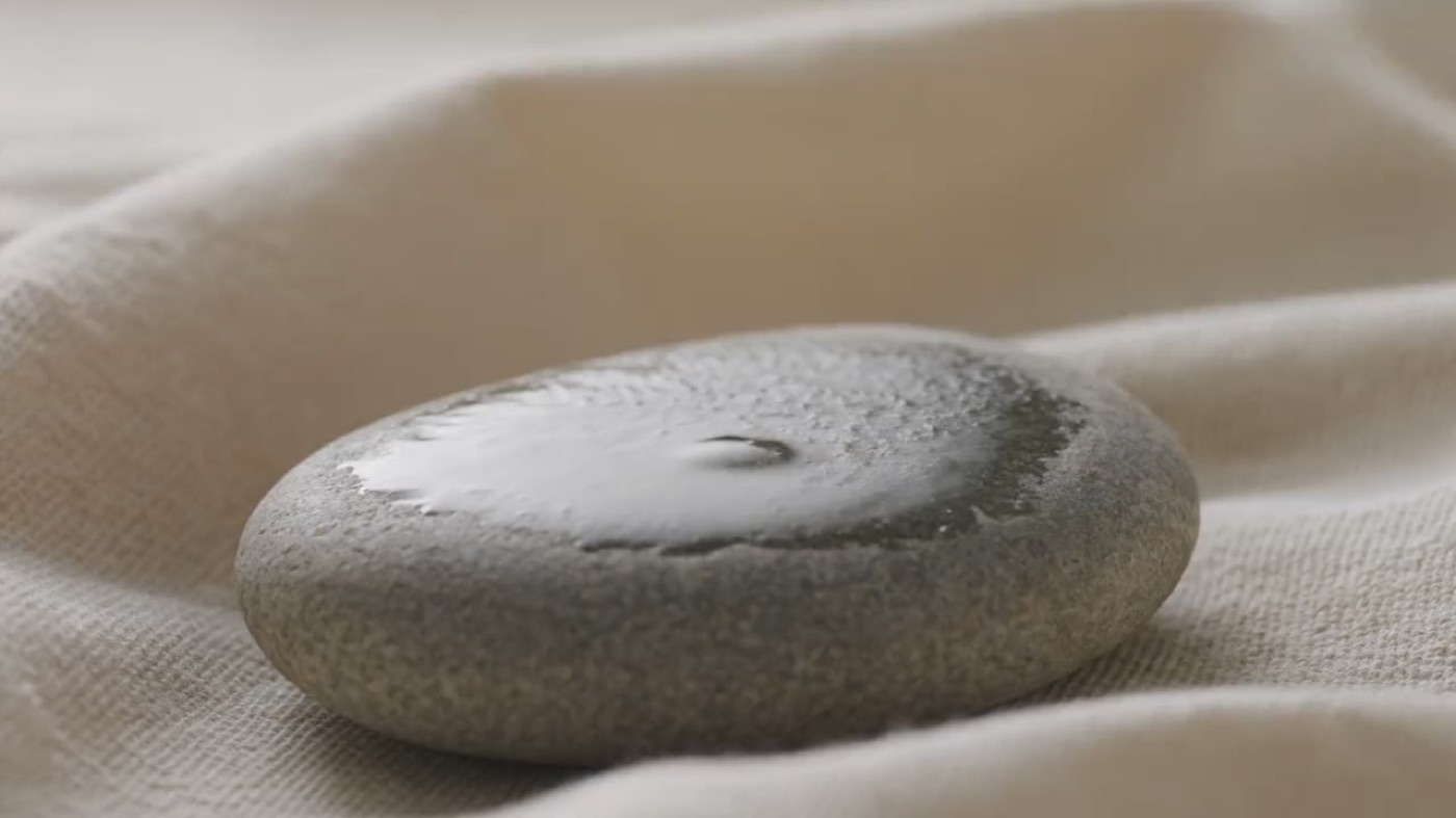 A drop of water falling onto a wet river stone, sending out one slow ripple.