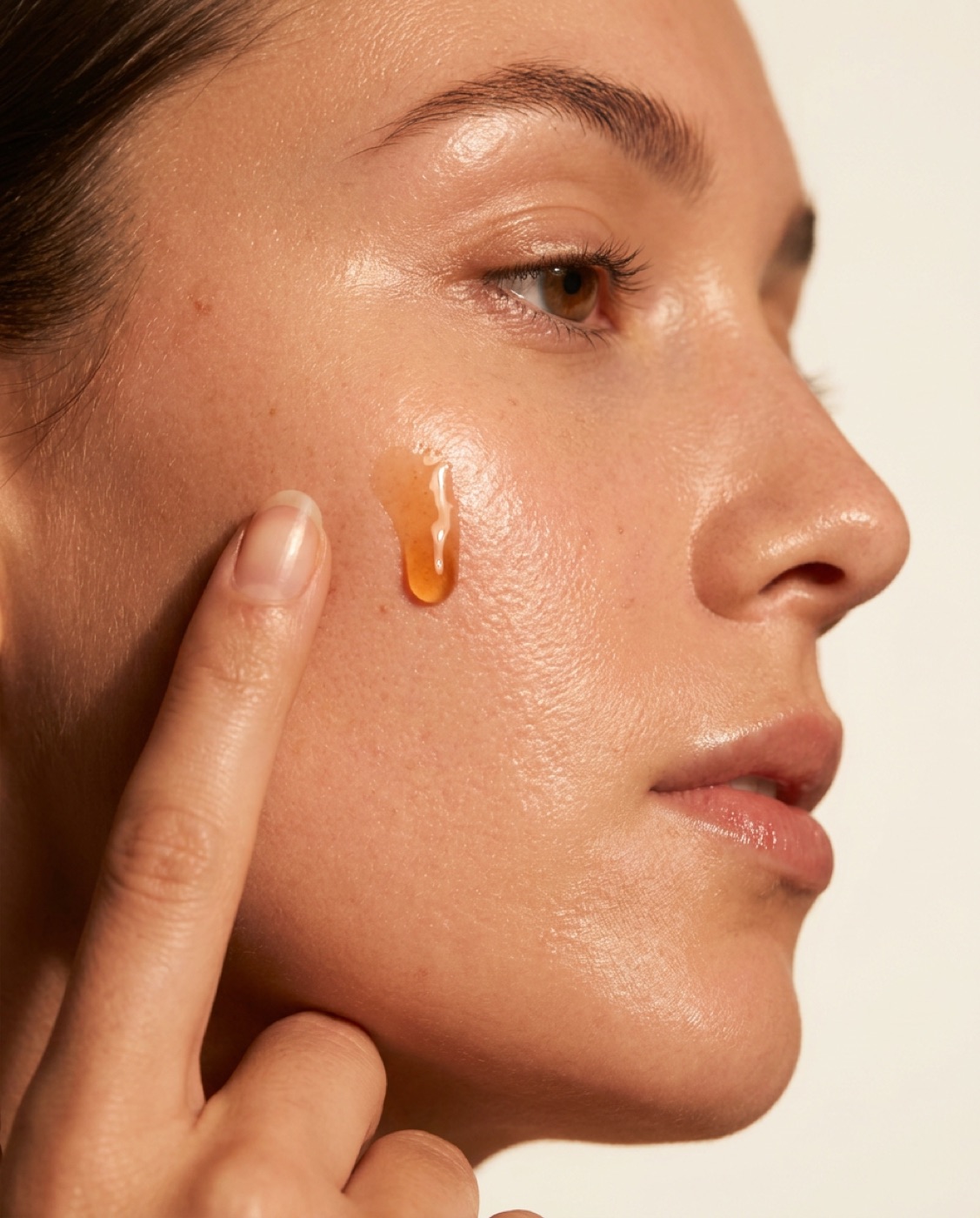 A drop of amber serum resting on a cheekbone, a fingertip about to press it in.