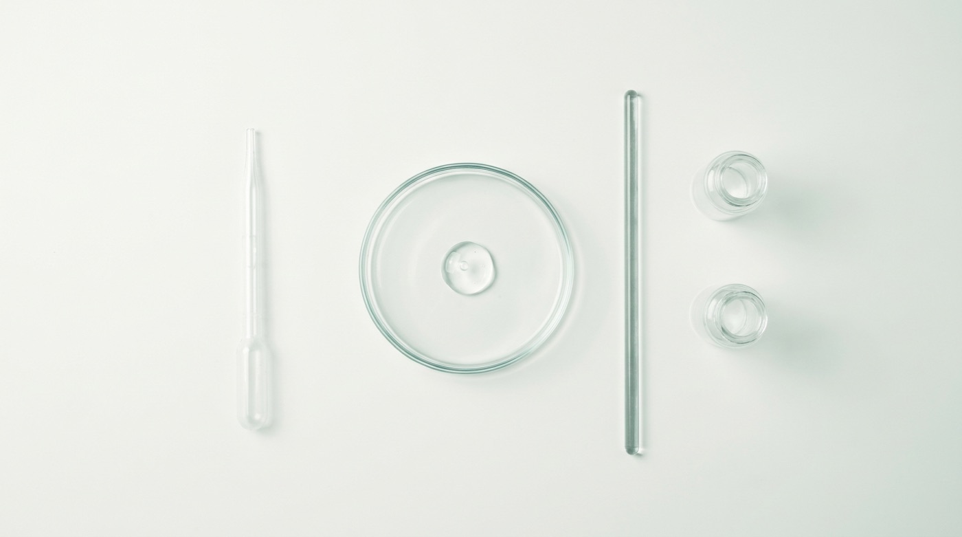 An overhead laboratory flat lay: pipette, petri dish of clear gel, stirring rod, and two glass vials on coconut-white.